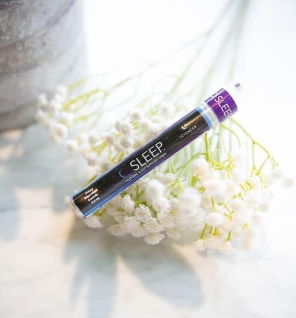 A spray bottle labeled 'SLEEP' positioned on a bed of white flowers, suggesting a calming or sleep-inducing product.